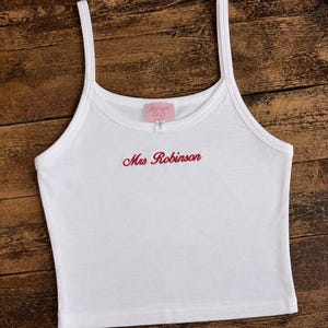 May include: A white sleeveless vest top with thin shoulder straps and a scoop neckline. The garment features the text Mrs Robinson embroidered in red cursive script across the chest and a pink brand label reading Romans Story at the neck.