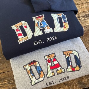May include: Navy and grey sweatshirts with the word "DAD" in a collegiate style, featuring teddy bear illustrations within the letters. The text "EST. 2025" is below the word "DAD". A navy sweatshirt has the name "Roman Gaia" embroidered on the sleeve.