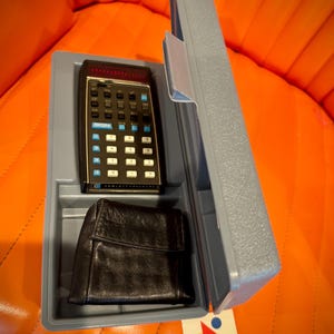 Könnte beinhalten: Ein Vintage-Taschenrechner von Hewlett-Packard mit einer schwarzen Ledertasche in einem grauen Kunststoff-Tragekoffer. Der Rechner hat eine schwarze Tastatur mit weißen und blauen Tasten und eine rote LED-Anzeige. Der Tragekoffer ist geöffnet.
