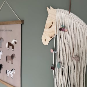 以下が含まれることがあります： 「Jonna」と刻まれた木製の馬の頭。馬の頭は、小さな木製のフルーツチャームが付いた白いマクラメの壁掛けに付けられています。