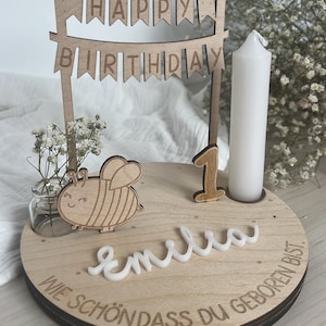 May include: Wooden birthday cake topper with a banner that reads "HAPPY BIRTHDAY". The topper includes a wooden bee, the number "1", a white candle, and the name "Emilia". The base has the phrase "WIE SCH&Ouml;N DASS DU GEBOREN BIST" engraved.