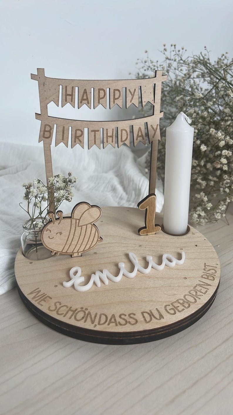May include: Wooden birthday cake topper with a banner that reads "HAPPY BIRTHDAY". The topper includes a wooden bee, the number "1", a white candle, and the name "Emilia". The base has the phrase "WIE SCH&Ouml;N DASS DU GEBOREN BIST" engraved.