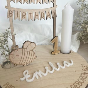 May include: Wooden birthday cake topper with a banner that reads "HAPPY BIRTHDAY". The cake topper includes a wooden bee, the number "1", a white candle, and the name "Emilia".