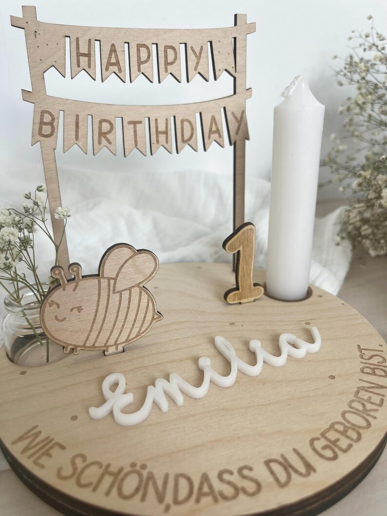 May include: Wooden birthday cake topper with a banner that reads "HAPPY BIRTHDAY". The cake topper includes a wooden bee, the number "1", a white candle, and the name "Emilia".