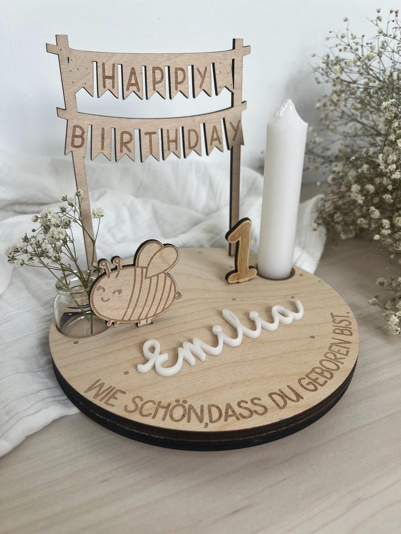 May include: Wooden birthday cake topper with a banner reading "HAPPY BIRTHDAY". The topper includes a candle, a wooden number 1, a bee, and the name "Emilia". The base has the text "WIE SCH&Ouml;N, DASS DU GEBOREN BIST".