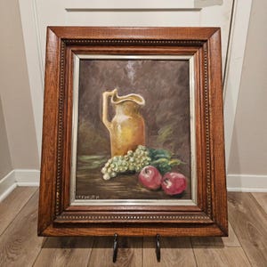 May include: An oil painting in a wooden frame depicts a still life. A yellow pitcher sits next to a bunch of green grapes and two red apples. The painting is signed "DEIKSEN" in the lower left corner.