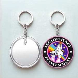 May include: Two keychains. One is a clear, round keychain. The other is a round keychain with a pink border and the text "EQUALITY IS NOT ROCKET SCIENCE". The keychain features a rocket ship, rainbow, and stars.