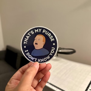 May include: A round sticker with a blue background and white border. The sticker features a cartoon image of a man with a surprised expression. The text on the sticker reads "That's my purse. I don't know you."