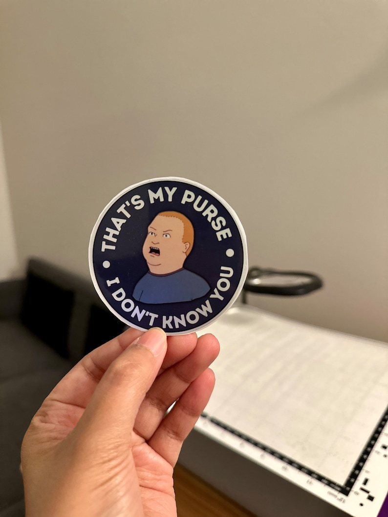 May include: A round sticker with a blue background and white border. The sticker features a cartoon image of a man with a surprised expression. The text on the sticker reads "That's my purse. I don't know you."