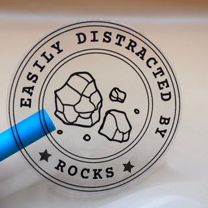 May include: A circular transparent sticker featuring a black line drawing of two rocks in the center. The text "EASILY DISTRACTED BY ROCKS" is printed in a circular border around the rocks, with two small black stars at the bottom.