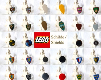 May include: A collection of 36 LEGO shields with various designs and colors, including a red, white, and blue shield with a lion, a green shield with a dragon, and a black shield with a skull. The shields are displayed on white LEGO minifigure bases. The text "LEGO" and "Schildes / Shields" is displayed in the center of the image.