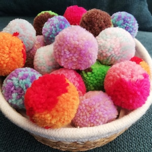 Könnte beinhalten: Ein Korb gefüllt mit bunten Pompons in verschiedenen Farbtönen von Rosa, Lila, Orange, Grün und Braun. Die Pompons sind aus Garn gefertigt und weich und flauschig.