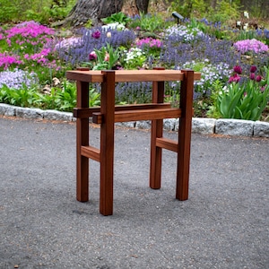 May include: A handcrafted, wooden side table with a rich, reddish-brown finish. The table features a rectangular top and a lower shelf, supported by a sturdy, geometric frame. The table stands approximately 30 inches tall.