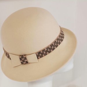 Può includere: Un cappello cloche in feltro color crema con un nastro a motivo marrone e crema e un fiocco.