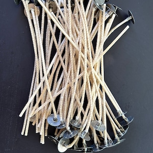 Könnte beinhalten: Ein Stapel naturfarbener Kerzen-Dochte mit Metallsockeln. Die Dochte sind gebündelt und die Metallsockel sind am unteren Ende des Stapels sichtbar.