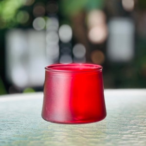 May include: A red glass candle holder with a frosted finish. The holder is round and has a wide base.