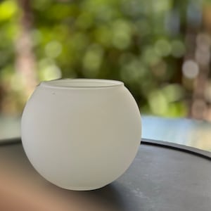 May include: A frosted glass, spherical vase. The vase has a smooth, rounded shape and a wide opening at the top. The glass has a soft, opaque appearance. The vase is sitting on a dark surface with a blurred green background.