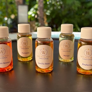 May include: Five small glass bottles with white lids and labels. The labels are white with a gold circle and the text "Wicks & Wax Candle Supplies". Each bottle contains a different colored liquid and has a different scent: Ocean Breeze, Pumpkin, Vanilla Bean, Mango, and Bayberry.