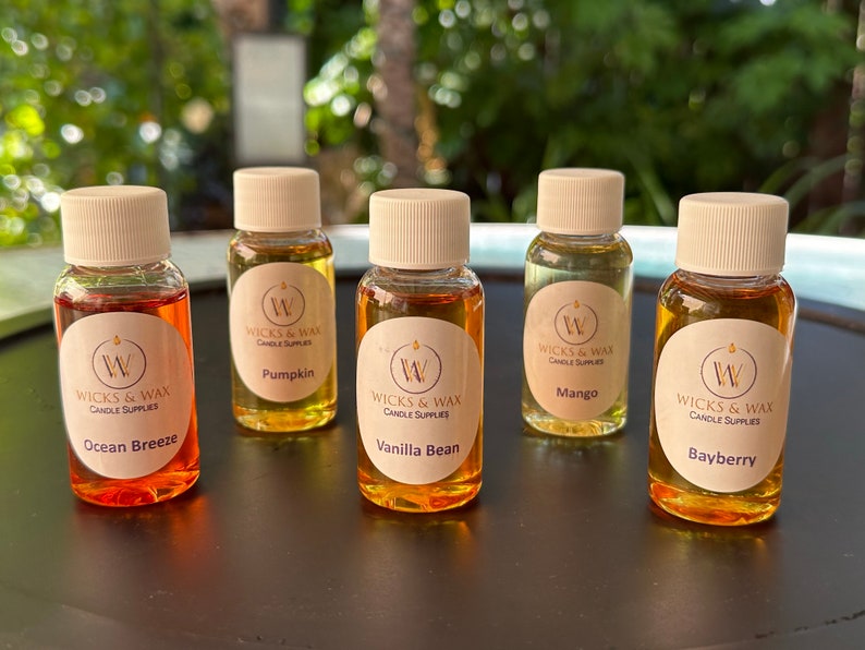 May include: Five small glass bottles with white lids and labels. The labels are white with a gold circle and the text "Wicks & Wax Candle Supplies". Each bottle contains a different colored liquid and has a different scent: Ocean Breeze, Pumpkin, Vanilla Bean, Mango, and Bayberry.