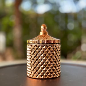 Könnte beinhalten: Ein goldener Kerzenbehälter mit geometrischem Muster und einem goldenen Deckel. Der Behälter steht auf einer braunen Oberfläche.