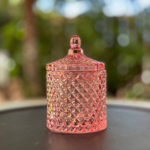 May include: A pink glass candle jar with a diamond pattern. The jar has a lid with a knob on top.
