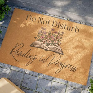 May include: A rectangular doormat with the text "Do Not Disturb Reading in Progress". The mat is tan with a graphic of an open book with flowers. The text is in a black, elegant font.