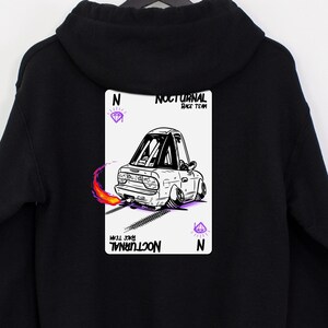 May include: Black hooded sweatshirt with a graphic of a cartoon car on a playing card. The card has the text "NOCTURNAL RACE TEAM" and a diamond symbol. The car is white with purple and orange flames.
