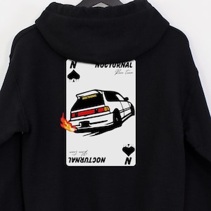 May include: Black hoodie featuring a car graphic on a playing card design. The card displays the word "NOCTURNAL" and a car with flames. The hoodie is suspended from a black hanger.