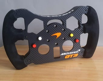 May include: Black racing steering wheel with a carbon fibre design and orange and white accents. The steering wheel has a McLaren GT3 logo.