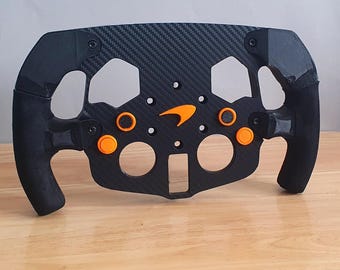 May include: A black racing steering wheel with orange buttons and a McLaren logo. The wheel is made of carbon fibre and has a textured surface.