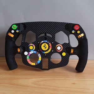 May include: A black racing steering wheel with a carbon fibre finish. It features a variety of buttons, switches, and dials, including a yellow and red Ferrari logo in the centre.