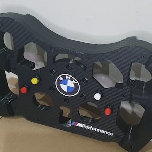 Könnte beinhalten: Schwarzes Carbonlenkrad mit BMW-Logo und M Performance-Branding. Das Lenkrad hat mehrere Knöpfe und einen weißen Knopf in der Mitte.