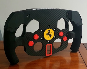Könnte beinhalten: Ein schwarzes Rennsportlenkrad mit Karbonfaser-Design und roten Knöpfen. Das Lenkrad hat ein gelbes Ferrari-Logo in der Mitte.