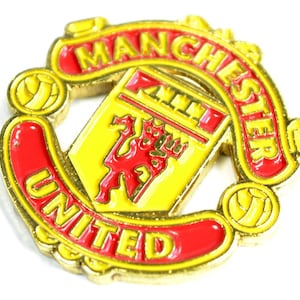 May include: A gold and red metal pin featuring the Manchester United logo. The logo depicts a red football, a red lion, and the words "Manchester United".