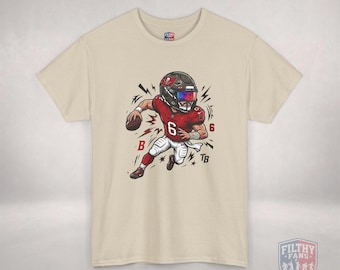 Puede incluir: Una camiseta beige de manga corta con una ilustración de dibujos animados de un jugador de fútbol americano con camiseta roja número 6, casco negro con visera tintada y pantalones blancos. El gráfico incluye detalles de rayos rojos y negros.