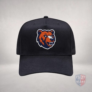 May include: A black baseball cap featuring an embroidered patch on the front panel. The patch displays a stylised bear head in shades of orange, dark blue, and white. The cap has a structured crown and a curved brim with visible stitching details.