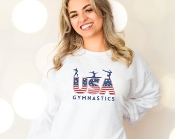 May include: White sweatshirt with a graphic of the word "USA" in red, white, and blue with stylized figures of gymnasts. The text "Gymnastics" is below the graphic.