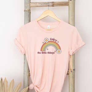 May include: A pink t-shirt with a rainbow and the text "Enjoy the little things" printed on the front. The rainbow is in shades of orange, yellow, green, blue, and purple. There are two white daisies on either side of the rainbow.