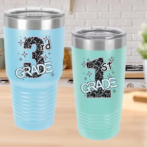 May include: Two insulated tumblers, one light blue and one mint green, with silver lids. The light blue tumbler displays "3rd GRADE" in a black, glittery font with star accents. The mint green tumbler shows "1st GRADE" in a similar style.