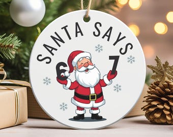 May include: A white Christmas ornament with the text "SANTA SAYS" and a cartoon Santa Claus holding up the numbers 6 and 7. The ornament has a string for hanging and is decorated with snowflakes.