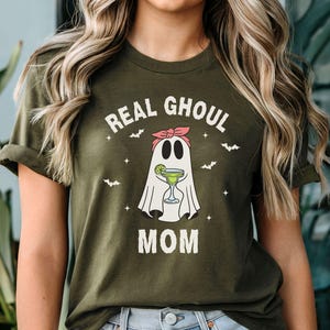 May include: Olive green t-shirt with the text "REAL GHOUL MOM" and a cartoon ghost holding a cocktail. The ghost wears a pink headband and is surrounded by bats and stars. A fun Halloween-themed graphic tee.
