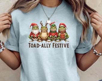 May include: Light blue t-shirt featuring four cartoon frogs dressed in Christmas attire. The frogs are wearing Santa hats and holding gifts. The text "TOAD-ALLY FESTIVE" is printed below the frogs.