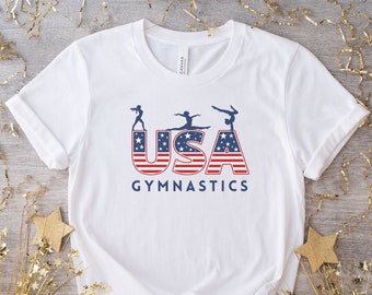May include: White t-shirt with a red, white, and blue USA graphic featuring three female gymnasts. The text "USA Gymnastics" is printed below the graphic.