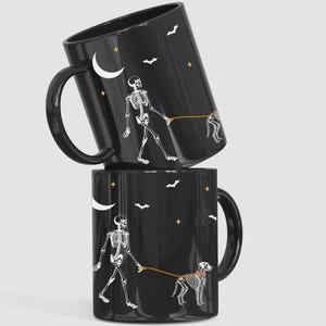 May include: Two black ceramic mugs stacked, each featuring a white skeleton walking a skeleton dog on a leash. The design includes a crescent moon, stars, and bats. The mugs have black handles.