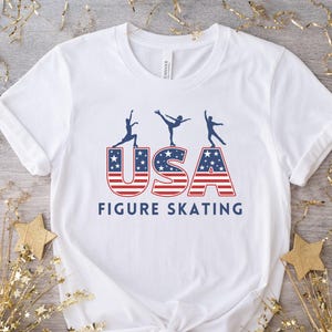A white t-shirt with a blue and red USA graphic with three figure skater silhouettes on top of the USA letters. The text ‘Figure Skating' is below the graphic.