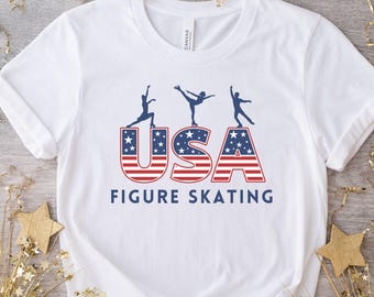 May include: White t-shirt with a USA Figure Skating design. The design features the word "USA" in red and blue stripes with stars, and three skater silhouettes above the text "FIGURE SKATING".