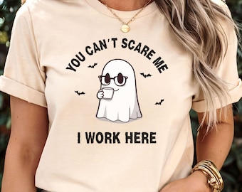 May include: Beige t-shirt with the text "YOU CAN'T SCARE ME I WORK HERE" and a cartoon ghost wearing glasses and holding a mug. The design includes small bat illustrations.