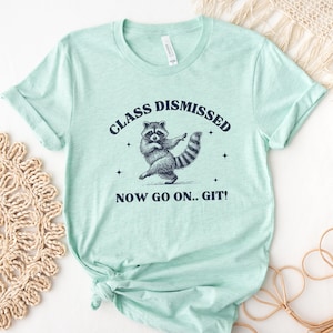 May include: A light green t-shirt with the text "CLASS DISMISSED" above a raccoon illustration. Below the raccoon is the text "NOW GO ON.. GIT!". The raccoon is depicted in a dancing pose.