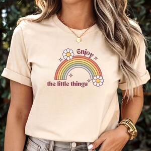 A woman wearing a natural colored t-shirt featuring a retro-style rainbow graphic with daisies and the text 'Enjoy the little things'. The shirt has short sleeves and a crew neck.  The design is in muted pastel colors, including orange, yellow, green, blue, and purple. The daisies are a creamy white with yellow centers. The text is in a maroon font. The overall aesthetic is vintage and positive.