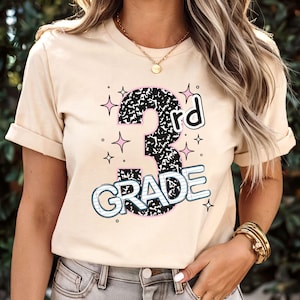 May include: Beige t-shirt with a colorful graphic for 3rd grade. The number 3 is black and white, outlined in pink, with the word "rd" in black. The word "GRADE" is in blue and white, with star accents.
